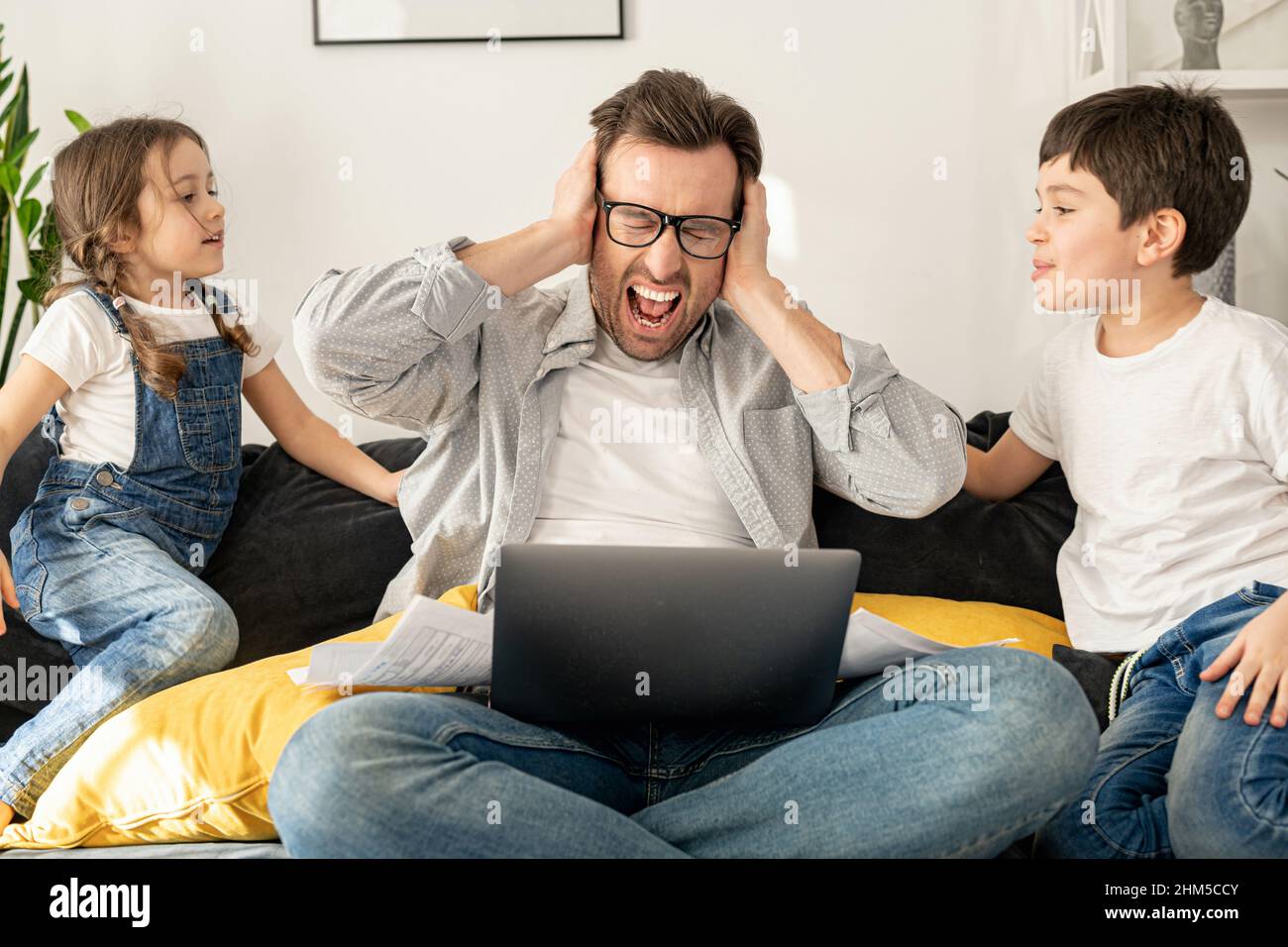 Pazza irritato padre urlando con gli occhi chiusi, chiudendo le orecchie con le palme, mentre i suoi due figli gridano insieme. Stress occupato singolo genitore è babysitting e lavoro a distanza Foto Stock