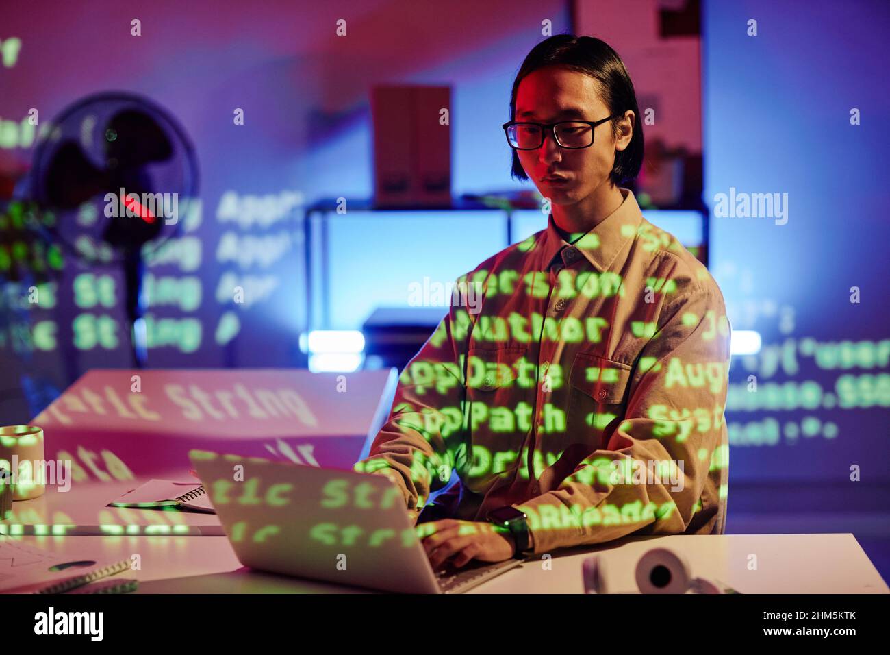 Giovane asiatico maschile diversità programmatore che lavora di fronte al computer portatile a tarda notte, mentre si siede da scrivania in moderno ufficio open space Foto Stock