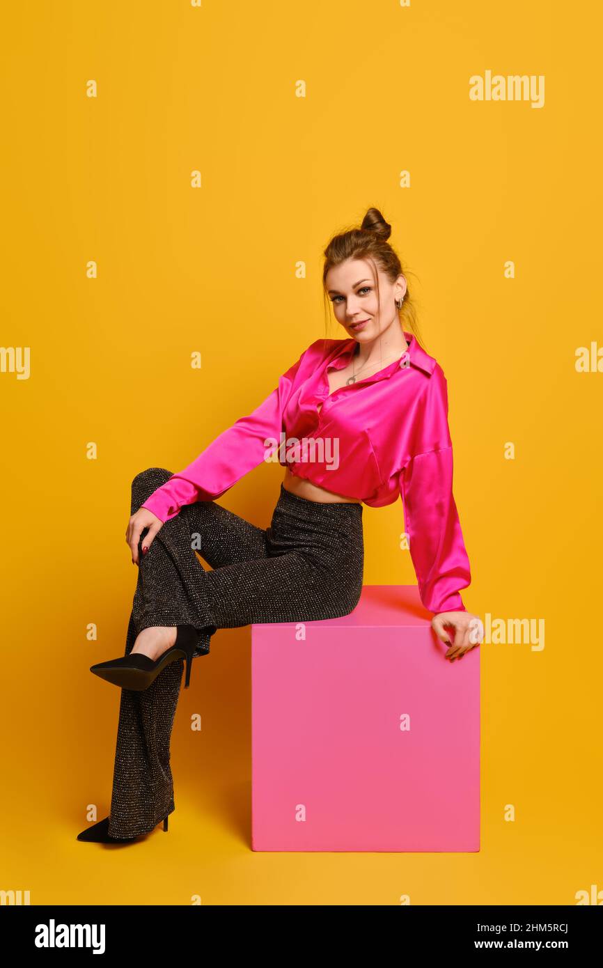 Donna giocosa in abiti vecchi e luminosi si siede in profilo su cubo viola su sfondo giallo luminoso studio Foto Stock