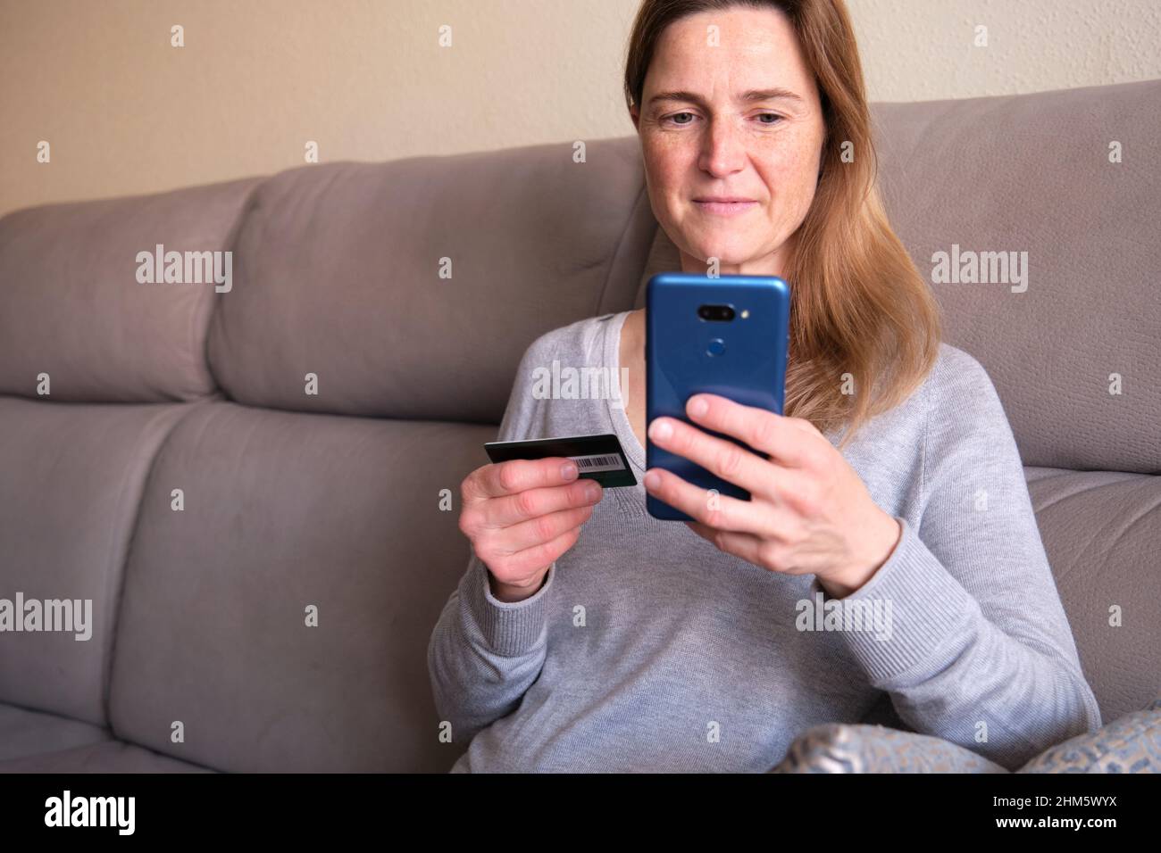 Donna felice che fa lo shopping online seduto sul divano a casa. Lady inserire il numero della carta di credito sul sito web con il suo smartphone. Foto Stock