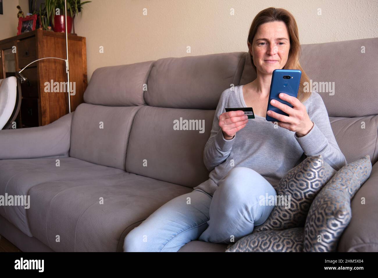 Donna felice che fa lo shopping online seduto sul divano a casa. Lady inserire il numero della carta di credito sul sito web con il suo smartphone. Foto Stock