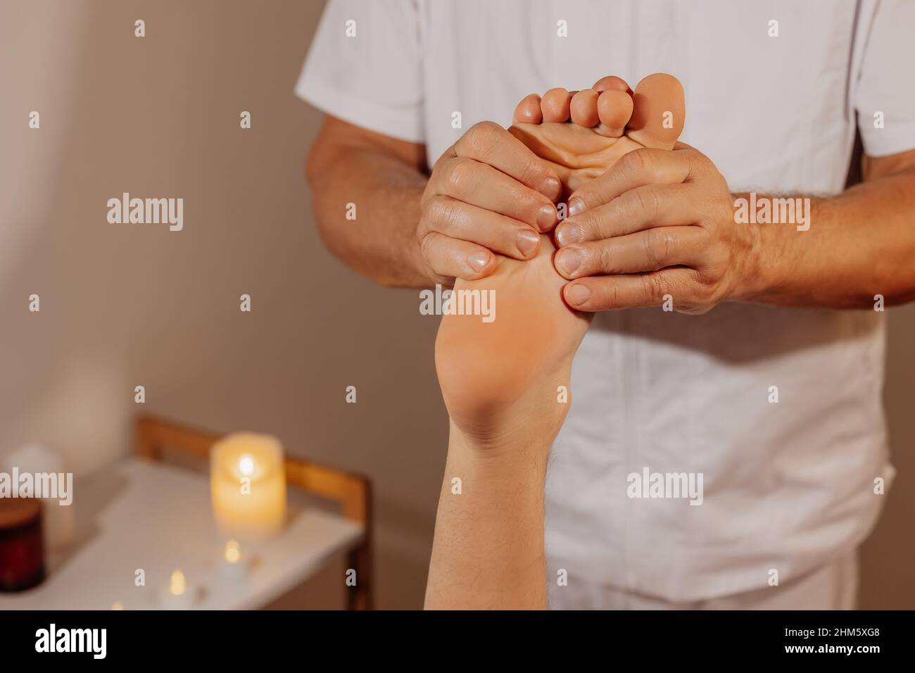 Massaggio piedi professionale nel salone SPA sullo sfondo delle candele. Vero massaggiatore in uniforme bianca che fa la terapia manuale per la gamba dell'uomo. Concetto di benessere, corpo e assistenza sanitaria. Foto Stock