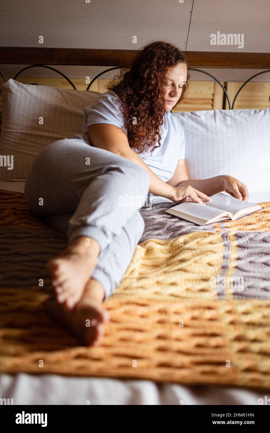 Donna seria bruna curly caucasica che riposa sul letto coperto di copriletto terry con il viso concentrato che tiene il libro in mani lettura casa Foto Stock