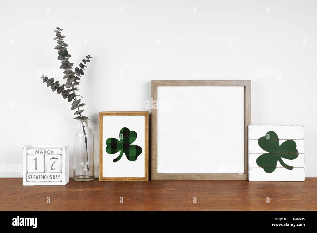 Mock up cornice in legno con St Patricks Day decor su una mensola in legno. Eleganti insegne in legno shabby, calendario e rami verdi. Cornice quadrata contro una parete bianca. Foto Stock