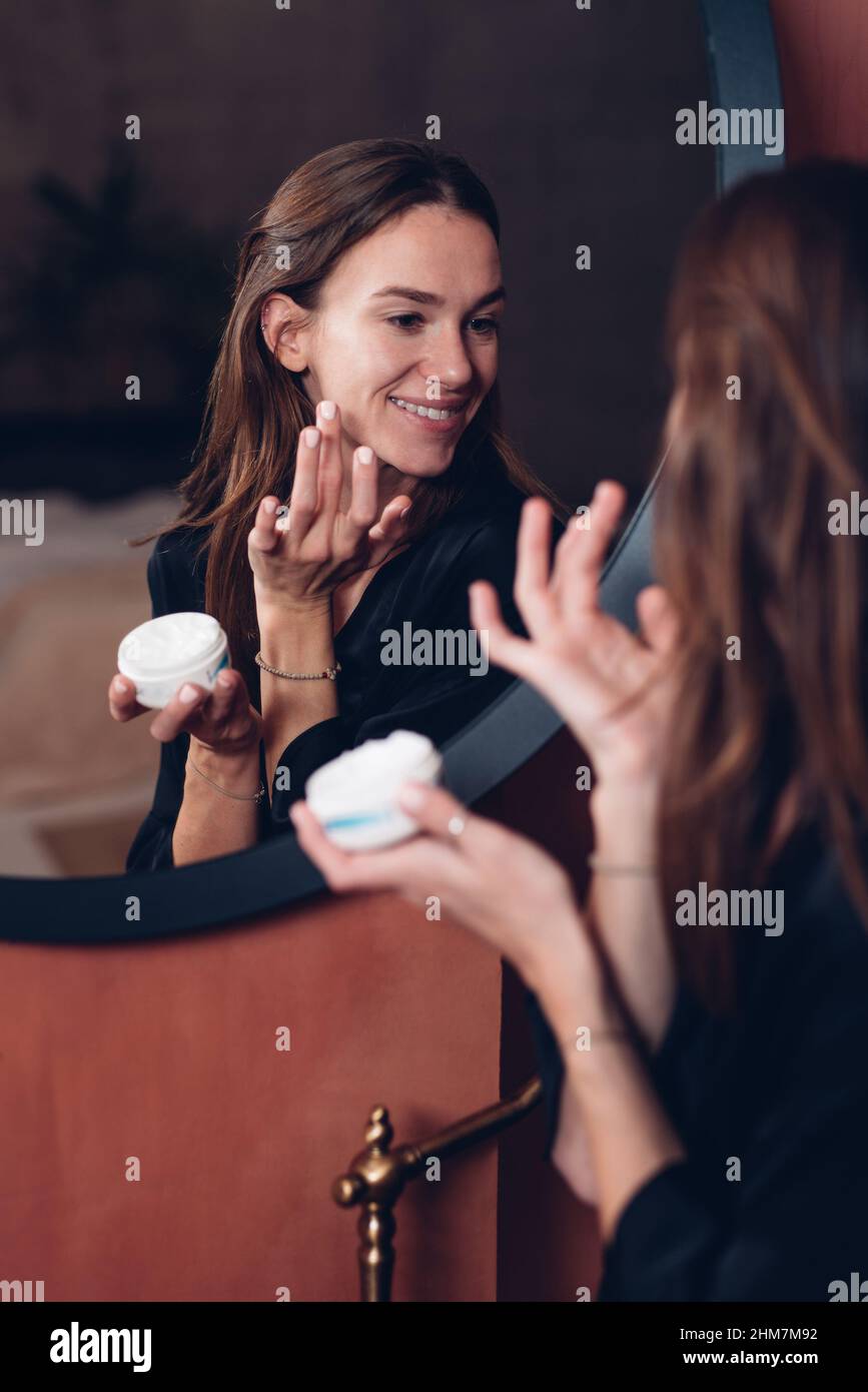 Giovane donna di fronte allo specchio che mette la crema sul suo viso. Foto Stock
