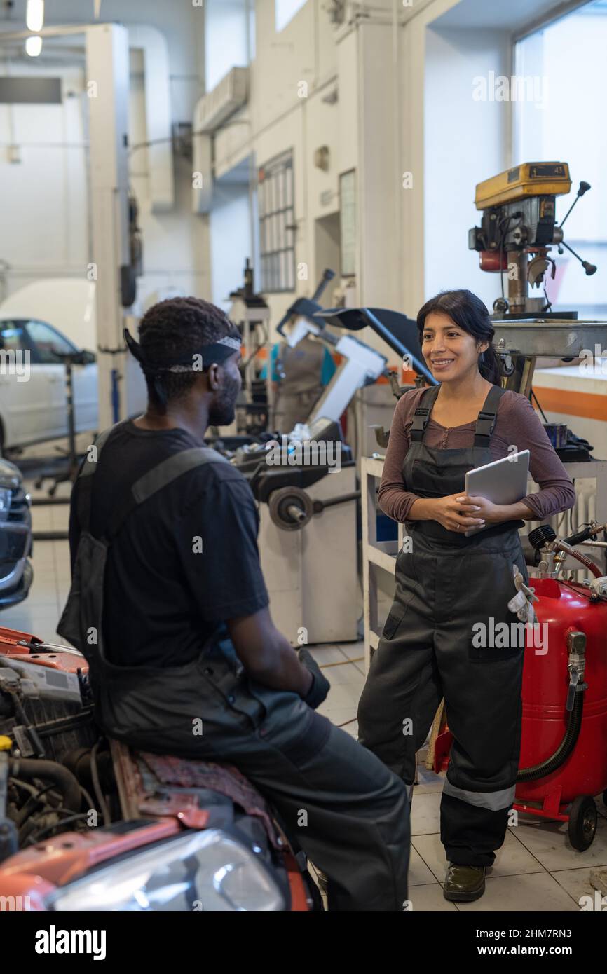 Ritratto verticale di due meccanici sorridenti dell'automobile che chiacchierano nel negozio del garage, fuoco sulla donna giovane nell'abbigliamento da lavoro Foto Stock