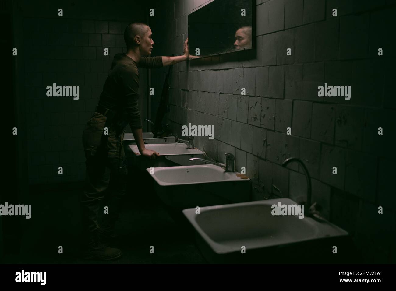 Vista laterale grandangolare per donna resistente con buzzcut guardando lo specchio in condizioni di sopravvivenza oscure, spazio di copia Foto Stock