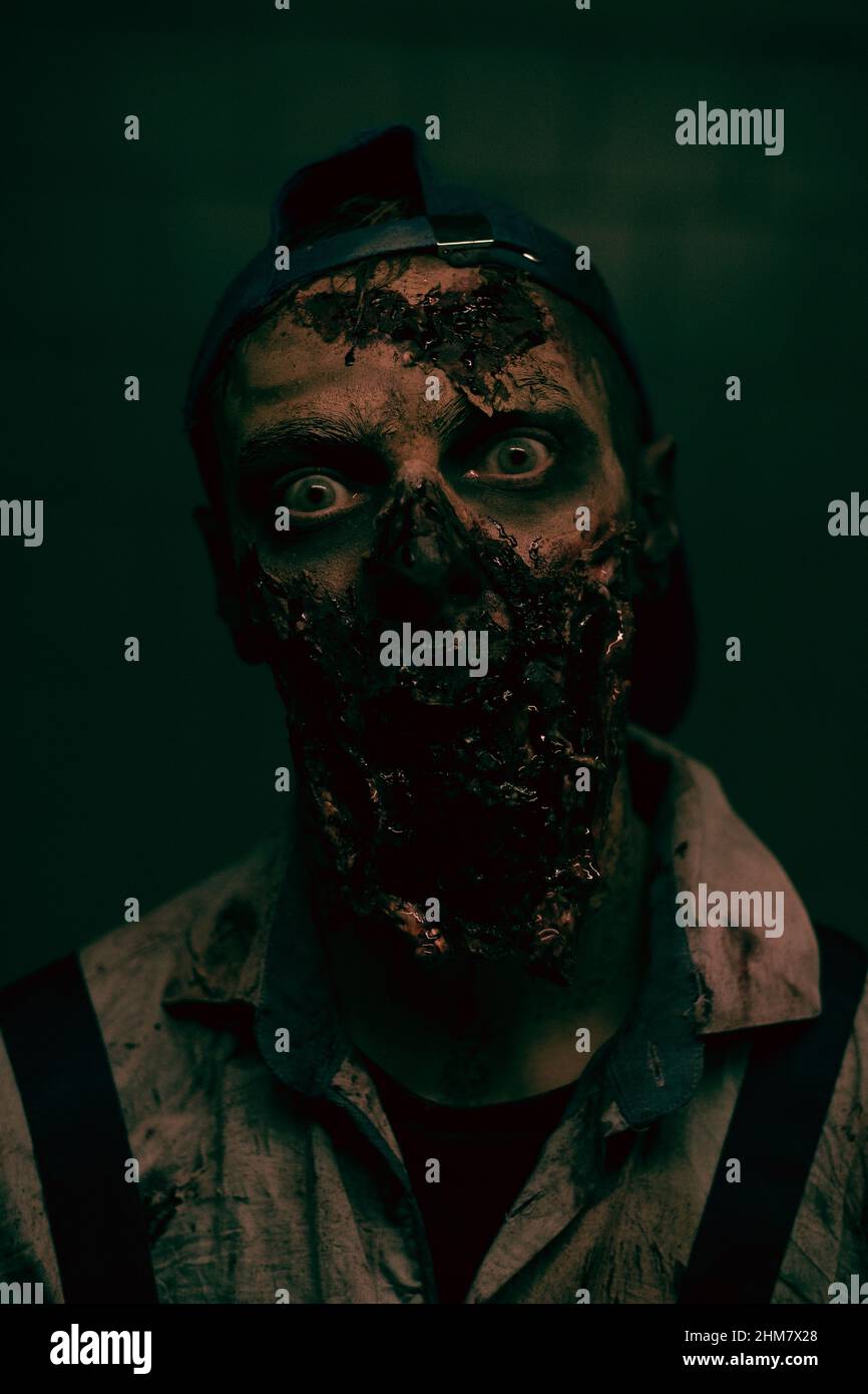 Primo piano verticale di zombie maschio creepy con volto sanguinoso guardando la fotocamera in oscuro ambiente horror Foto Stock