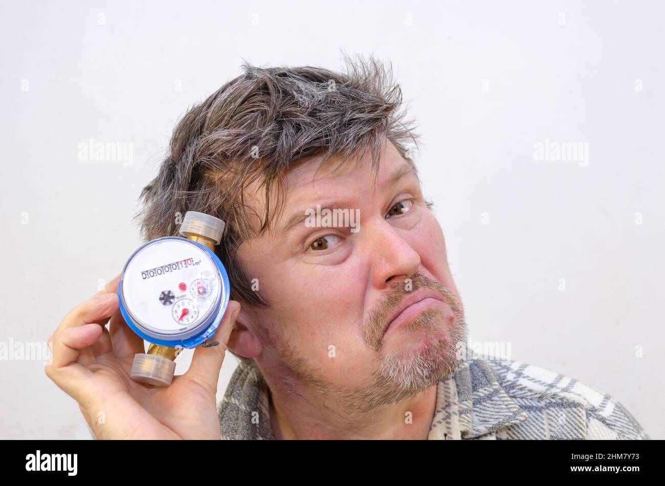 Il maschio grida e ascolta il contatore dell'acqua con esso all'orecchio. Uomo con capelli aggrovigliati e diseveleati davanti ad una parete di colore grigio chiaro. All'interno. Selez Foto Stock