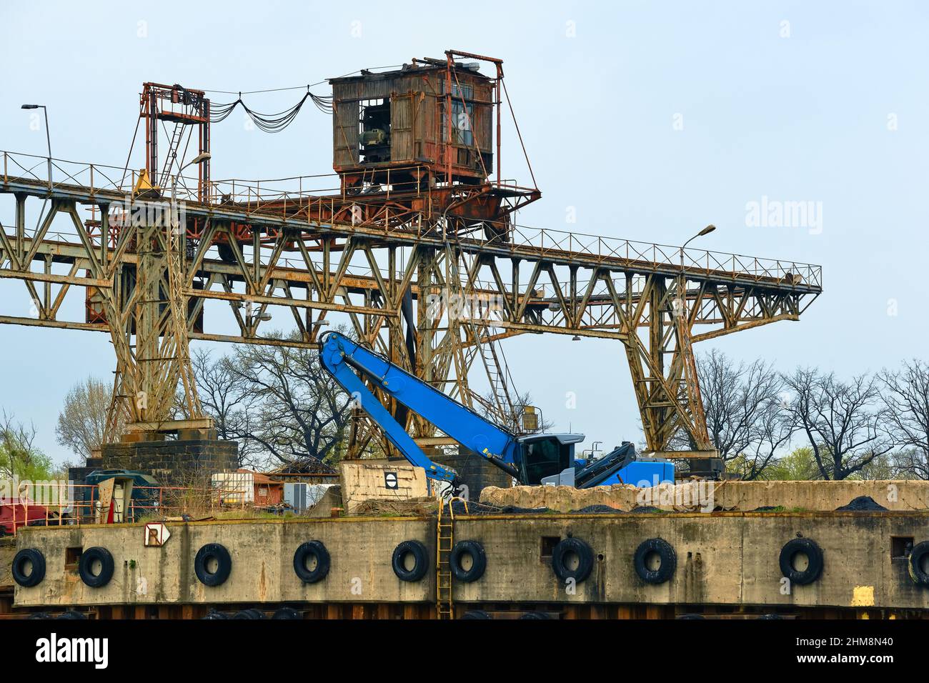 Vecchia gru a gantry e caricatore retroescavatore che lavorano sopra i detriti di costruzione nel porto del fiume in giorno coperto. Foto Stock