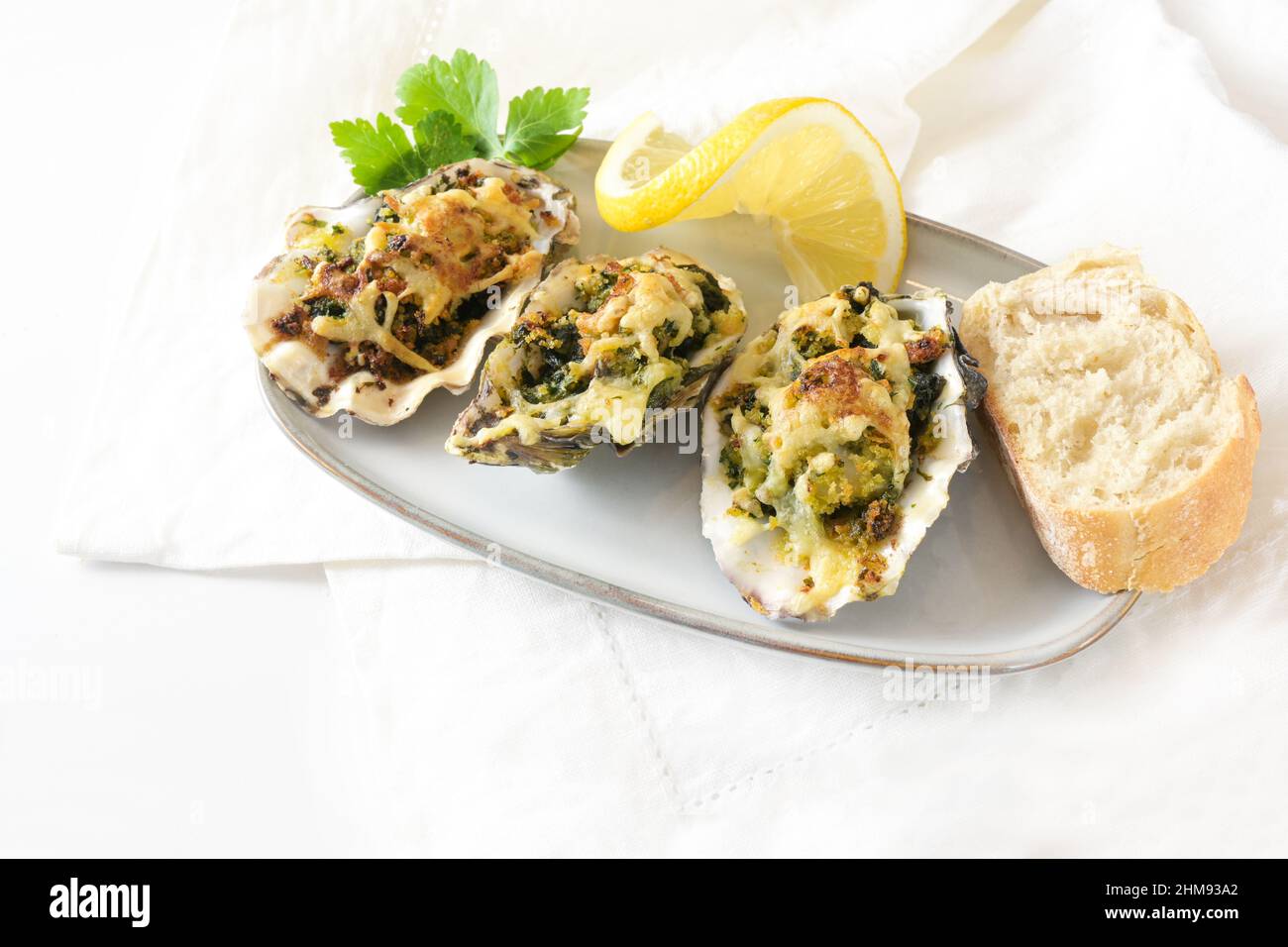 Tre ostriche al forno Rockefeller con spinaci e formaggio su un piccolo piatto con limone, pane e prezzemolo guarnire, aperitivo festivo su un tavolo bianco, co Foto Stock