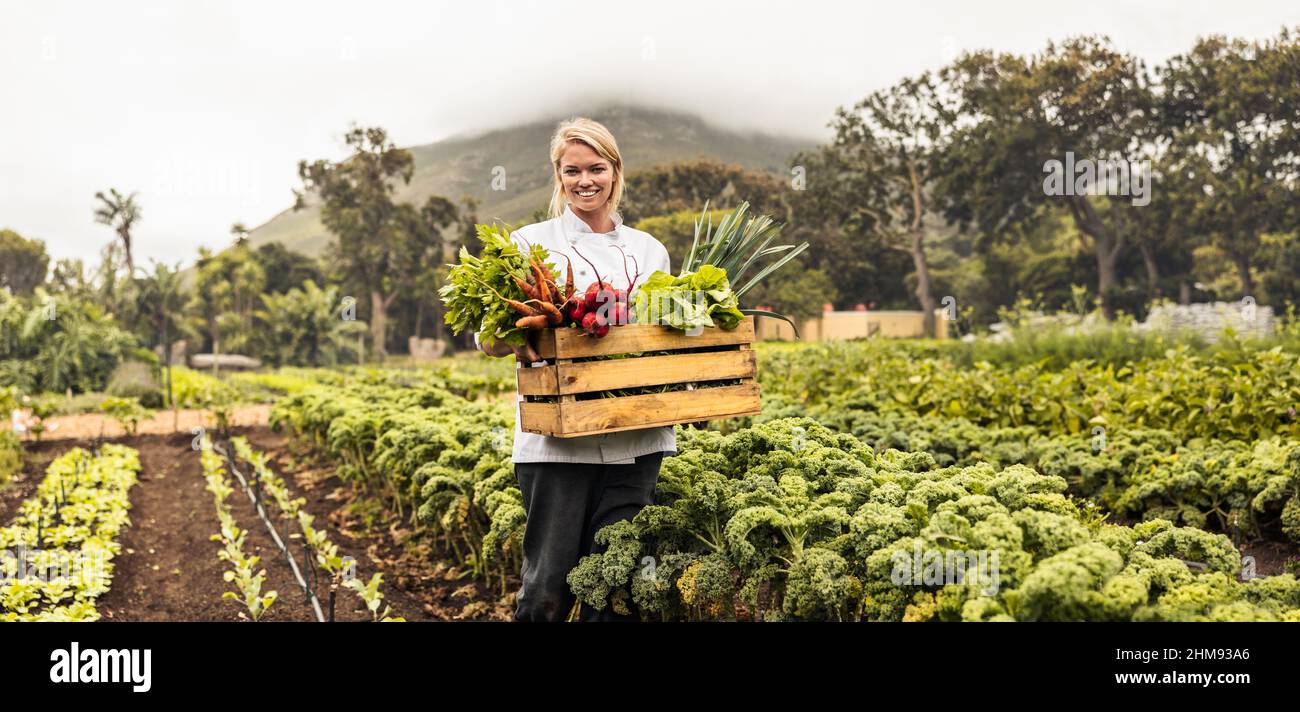 Trasporto di verdure fresche da fattoria a tavola. Allegro giovane chef femmina sorridente alla macchina fotografica mentre porta una cassa piena di verdure appena raccolte Foto Stock