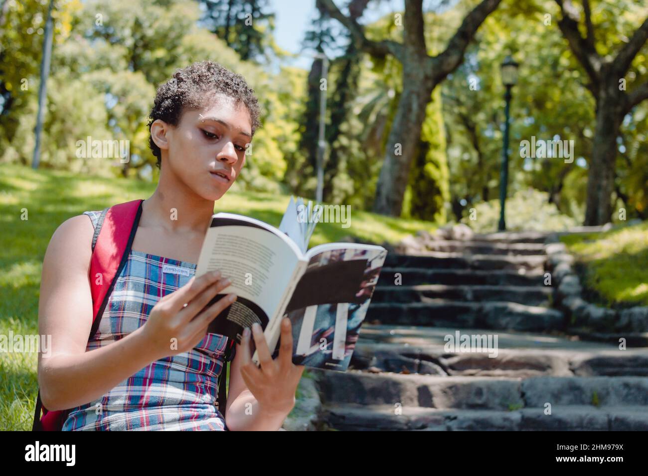 vista frontale della giovane bruna ispanica ragazza latina seduta sulle scale in un parco con alberi dietro di lei, lettura e studio con spazio copia, giorno libro Foto Stock
