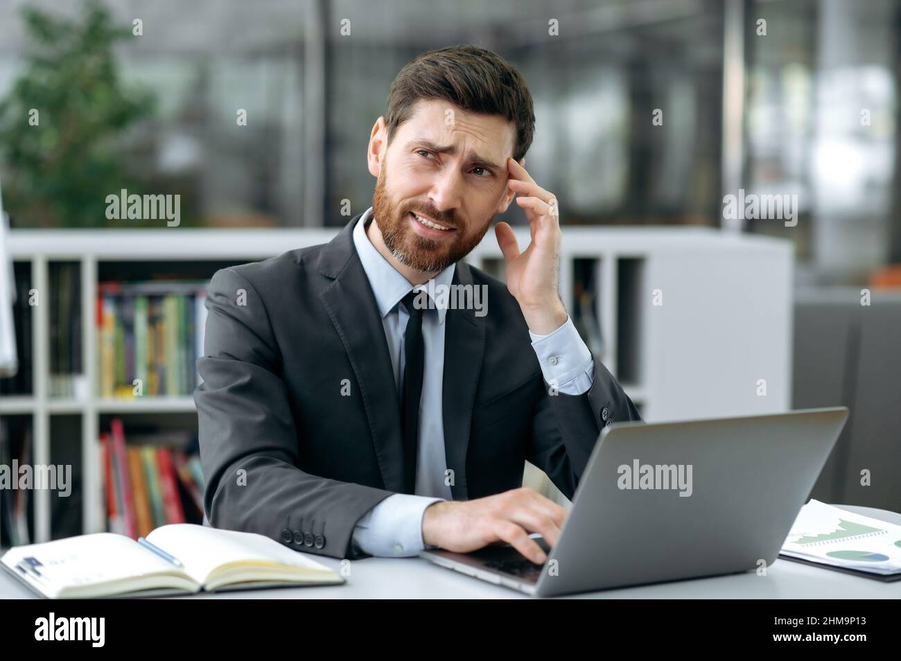 Triste sopravlavorato caucasico uomo d'affari, manager o proprietario di azienda, in ufficio moderno, che soffre di mal di testa, emicrania, rubs tempio, si sente stressato, stanco da lavoro straordinario, situazione stressante Foto Stock