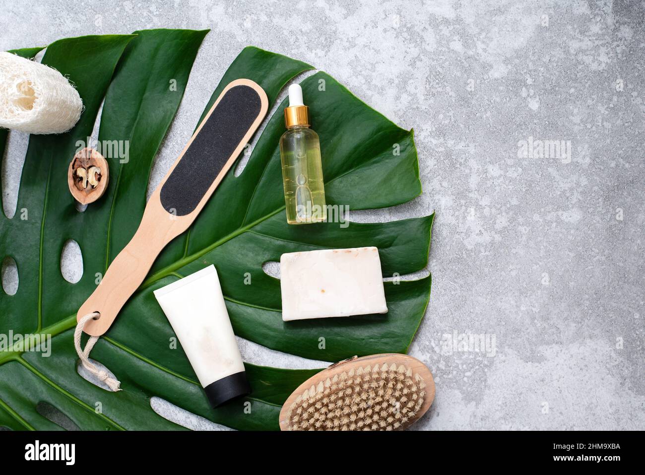 Set di prodotti cosmetici per la cura della pelle e del corpo femminile. Prodotti naturali biologici di bellezza spa, spazzola massaggio e foglie di monstera tropicale su grigio Foto Stock