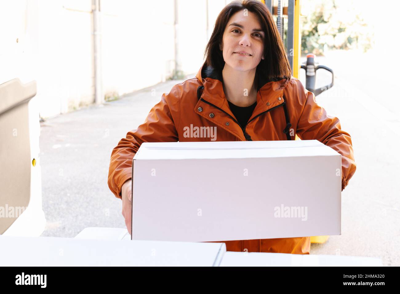 Contenuto donna spedizioniere lavoratore guardando la macchina fotografica mentre il carico pesante scatola bianca in camion parcheggiato su strada con impilatore professionale Foto Stock