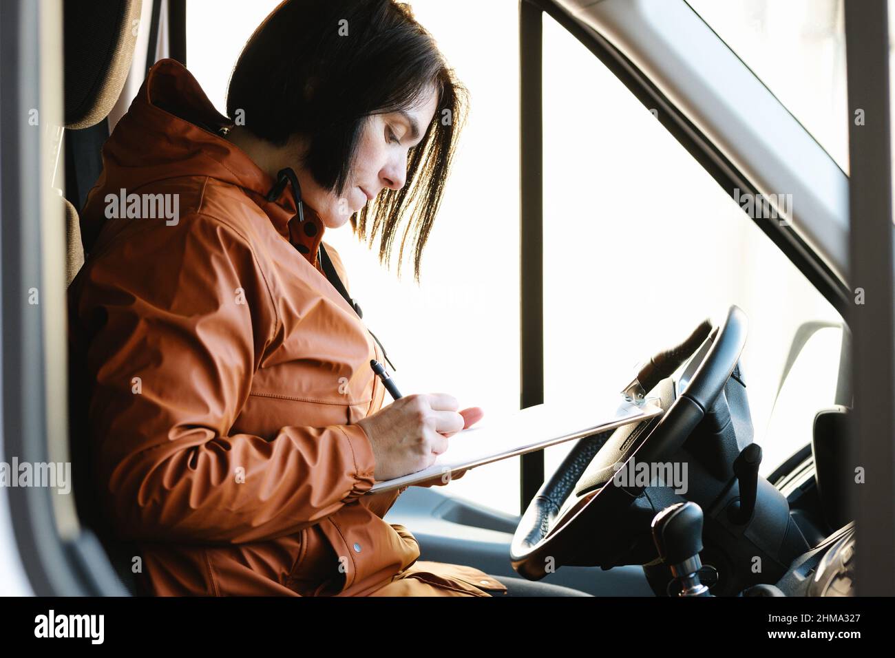 Vista laterale di una donna seria che scrive le note sulla clipboard mentre si siede in cabina del veicolo parcheggiato sulla strada durante il lavoro Foto Stock