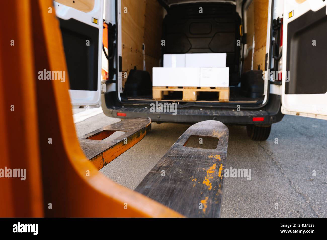 Impilatore industriale in metallo con forche regolabili poste su percorso asfaltato vicino al carrello aperto con impilamento di scatole di cartone su strada Foto Stock