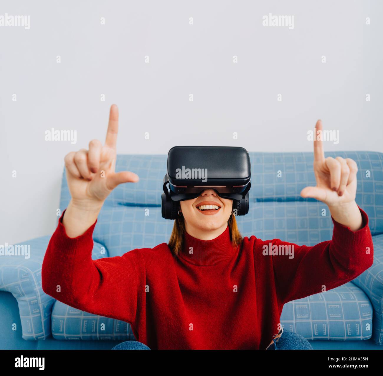Donna con occhiali di protezione per la realtà virtuale sorridente e rivolta verso l'alto mentre si siede sul divano ed esplora la realtà virtuale Foto Stock