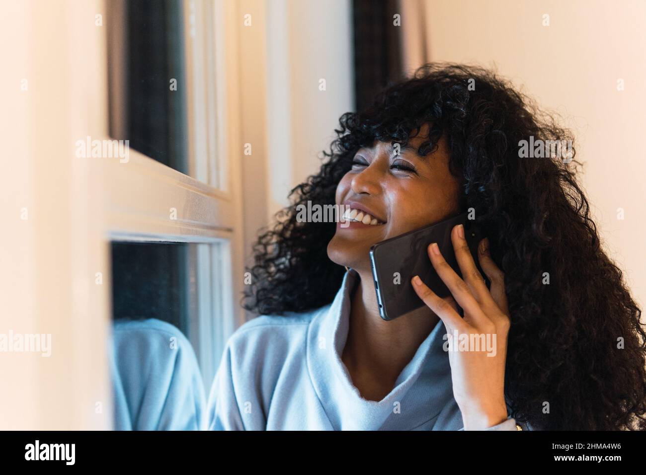 Giovane ragazza afroamericana allegra con capelli ricci scuri in elegante vestito caldo che parla telefono cellulare mentre guarda finestra di lancio Foto Stock