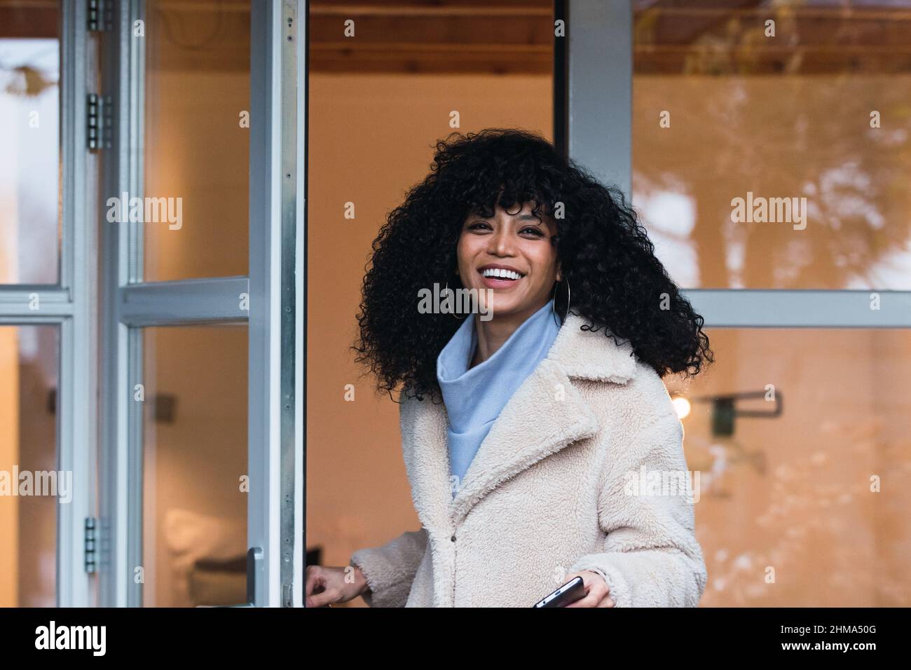 Giovane donna Africana americana allegra con capelli ricci scuri in un cappotto caldo che esce dalla casa attraverso la porta di vetro e guardando la macchina fotografica Foto Stock