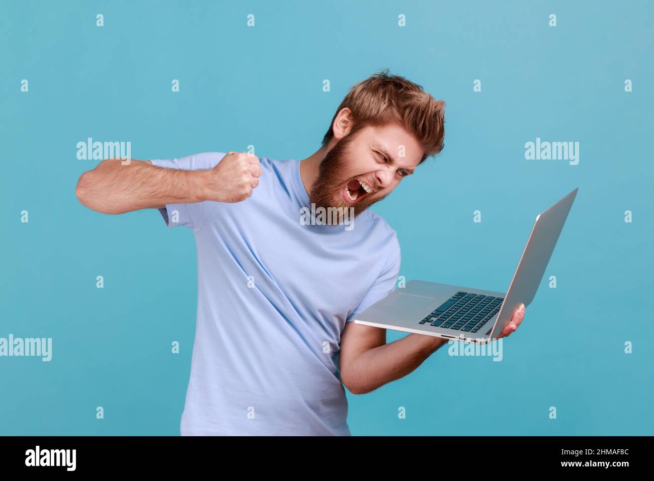 Ritratto di uomo che punzonano lo schermo del laptop, guardando con furious pazza espressione, boxe minacciando di colpire durante la conversazione in linea, videochiamata. Studio interno girato isolato su sfondo blu. Foto Stock