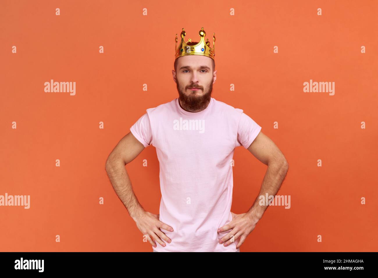 Ritratto di uomo bearded egoisticamente guardando la macchina fotografica, posando con la corona sulla testa, fingendo di essere re, stato privilegiato, indossando la T-shirt rosa. Studio interno girato isolato su sfondo arancione. Foto Stock