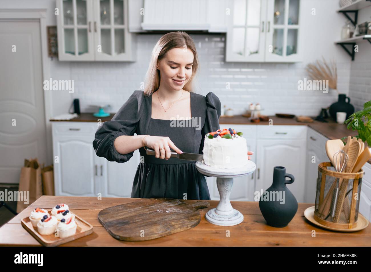 Pasticciere pasticciere giovane donna caucasica in abito grigio con coltello tagliato fetta di torta sul tavolo da cucina. Cakes cupcakes e dolce dessert monolocale in stile scandinavo Foto Stock