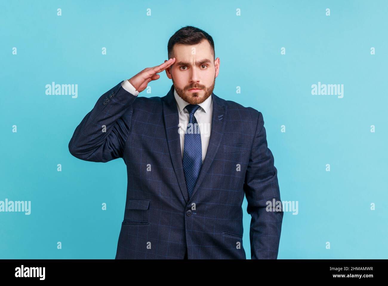 Sì Signore. Uomo d'affari serio con barba che indossa tuta ufficiale in stile che tiene la mano vicino alla testa, salendo, onore e gloria, militarizzazione. Studio interno girato isolato su sfondo blu. Foto Stock