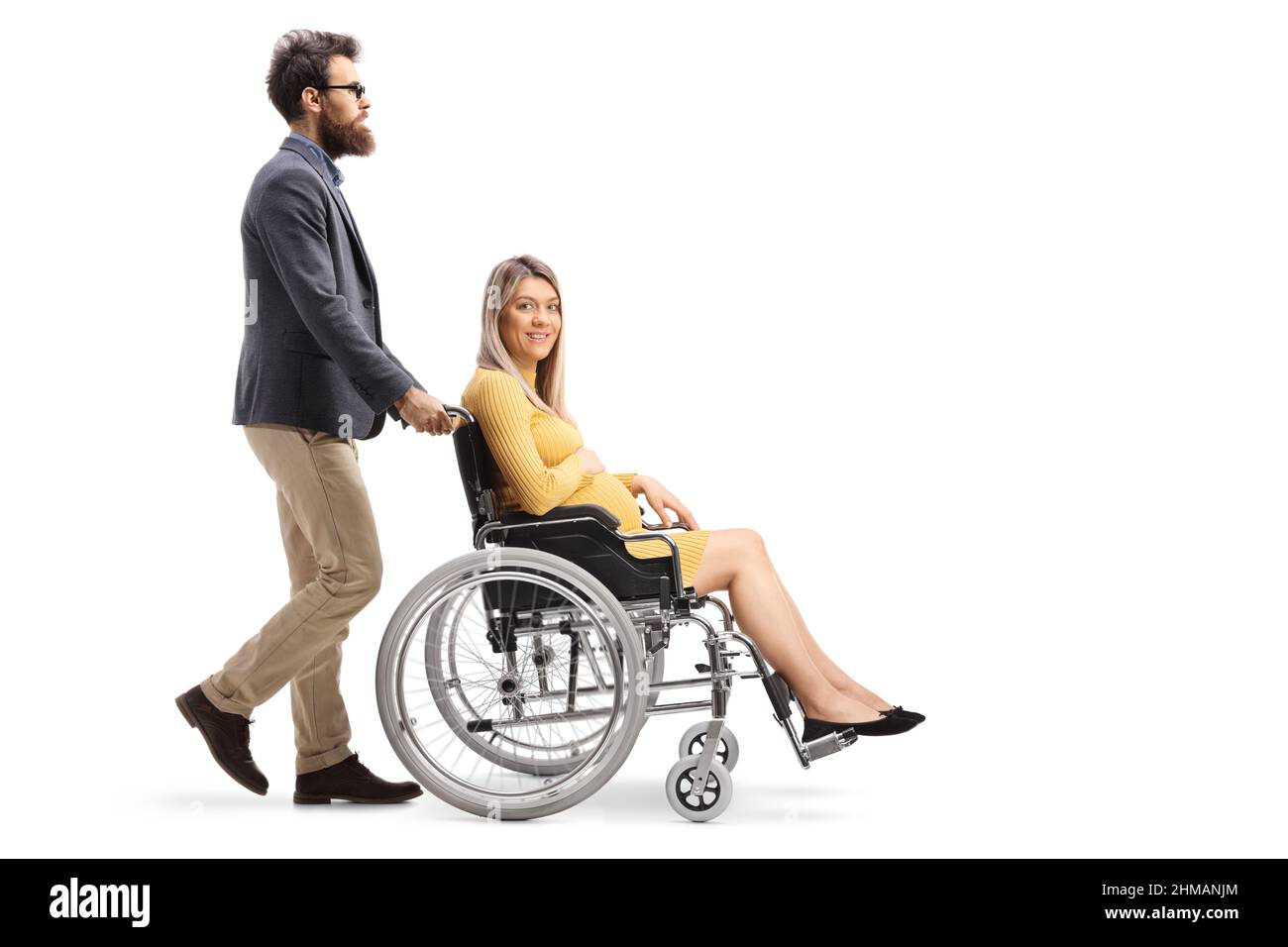 Colpo di profilo a tutta lunghezza di un uomo barbuto che spinge una donna incinta in una sedia a rotelle isolata su sfondo bianco Foto Stock