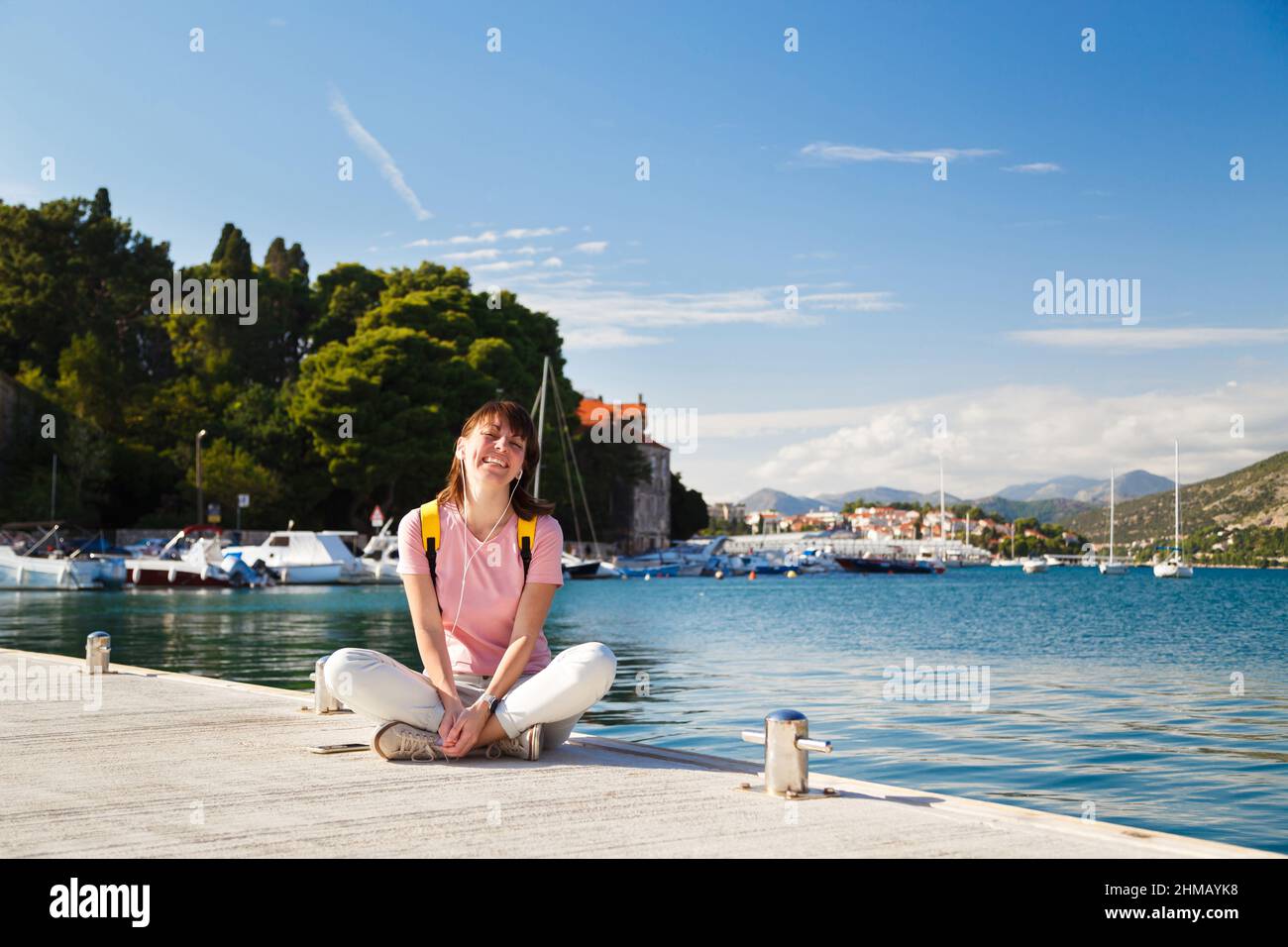 Giovane donna sorridente seduto a gambe incrociate sul molo, ascoltando musica con la costa croata sullo sfondo. Giornata di sole a Dubrovnik, Croazia. Concetto di stile di vita di viaggio Foto Stock