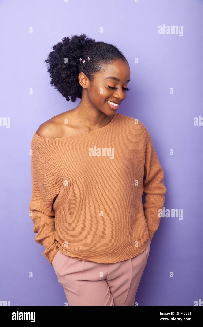 Una giovane donna nera in piedi vicino ad uno sfondo con le mani nelle sue tasche. Foto Stock