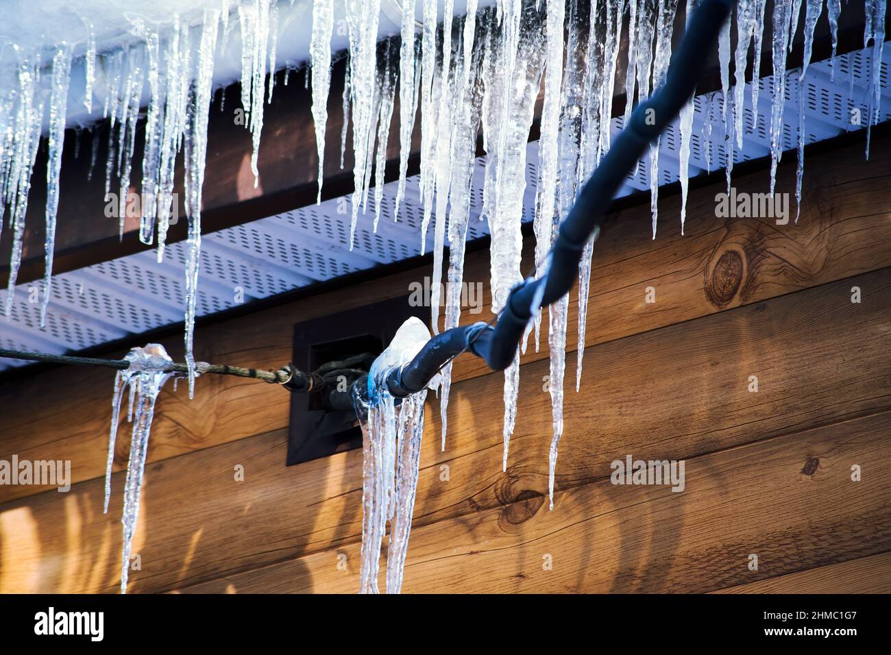 Le ghiacciole pendono dal tetto e dai fili elettrici in primavera in primi raggi di sole. Condizioni meteorologiche pericolose Foto Stock