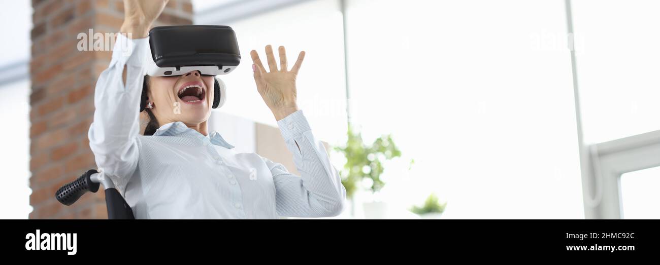 Donna seduta in sedia a rotelle indossando occhiali di realtà virtuale e agitando le mani Foto Stock