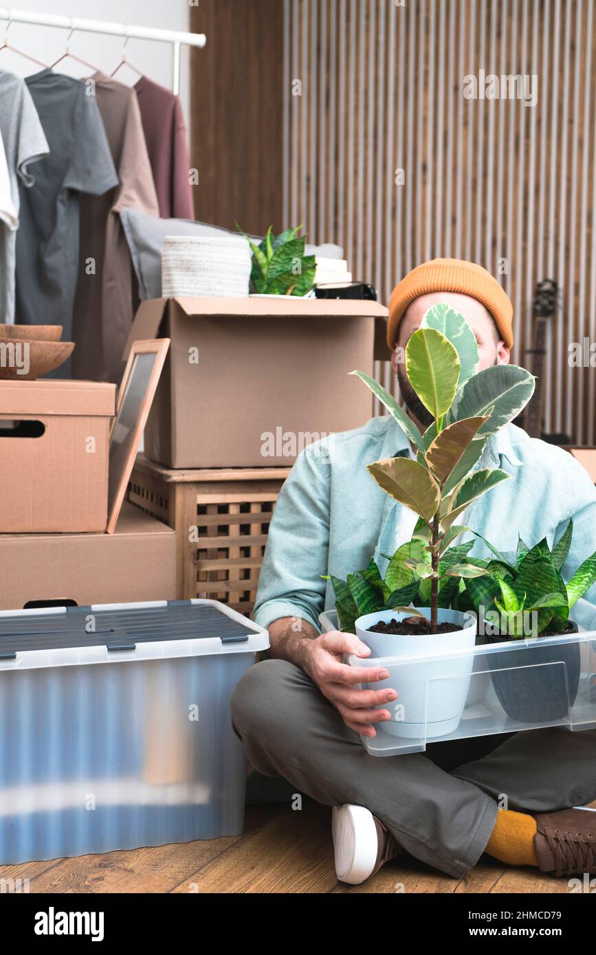 Un uomo maturo seduto in una casa non arredata, tenendo una scatola con piante. Un nuovo concetto di casa in movimento. Foto Stock