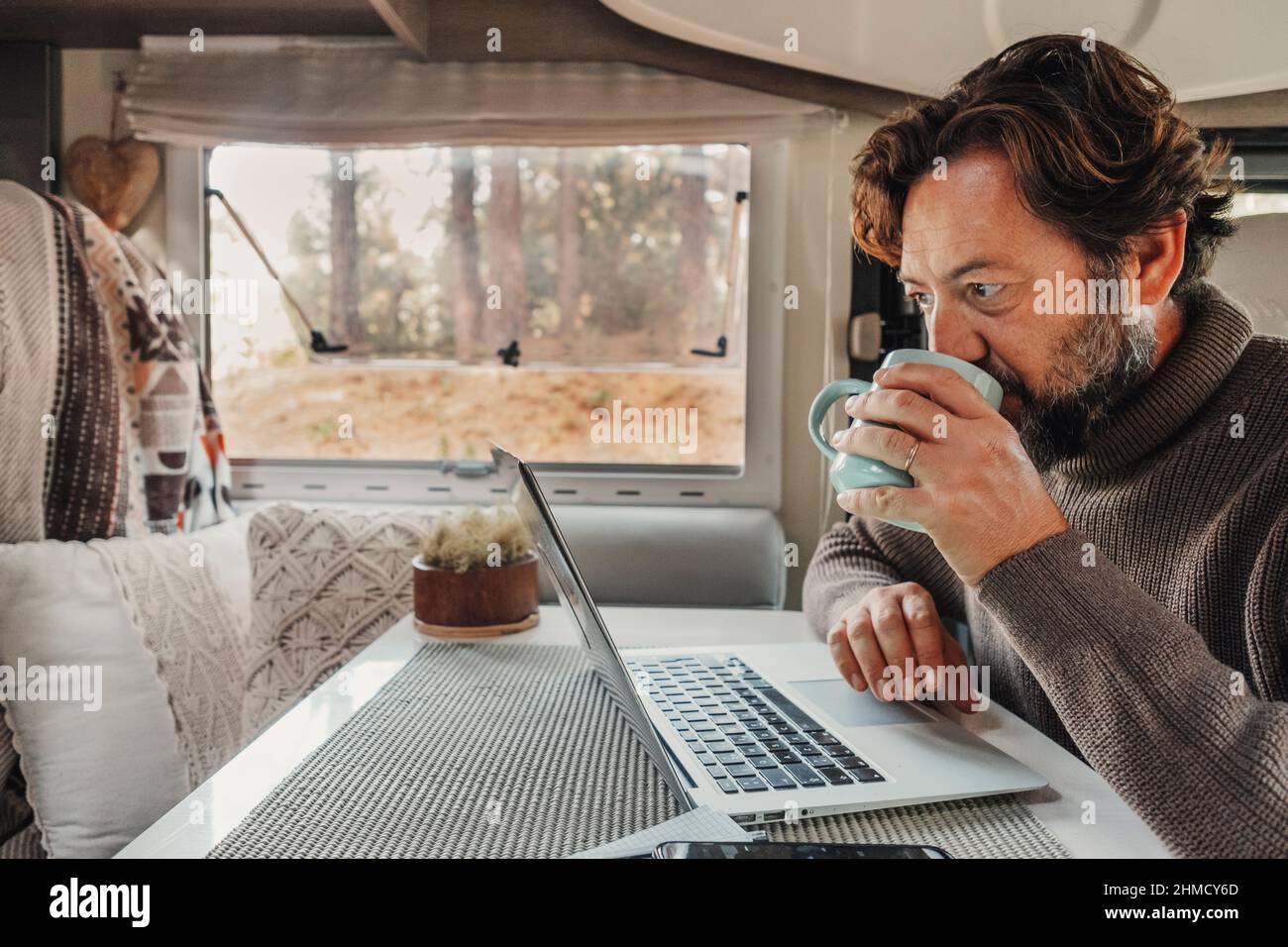 Uomo maturo che lavora su un computer portatile all'interno di un camper con la natura all'aperto vista fuori dalla finestra. Concetto di libertà e stile di vita all'avanguardia. Smar Foto Stock