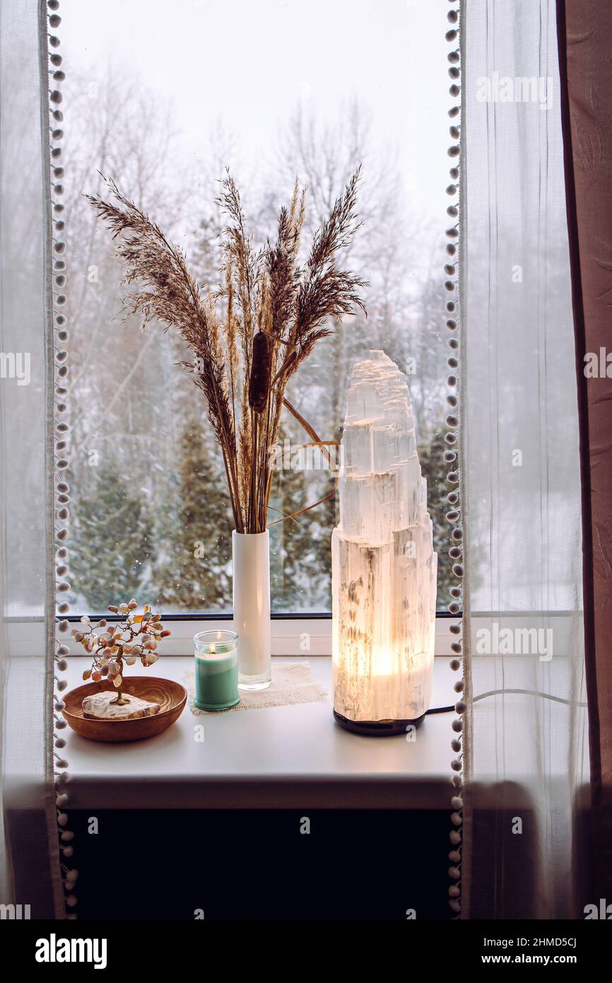 Ruvida grande selenite torre palo lampada illuminata sulla soglia della finestra di casa, spirituale decorazione accento casa. Inverno foresta sullo sfondo. Foto Stock