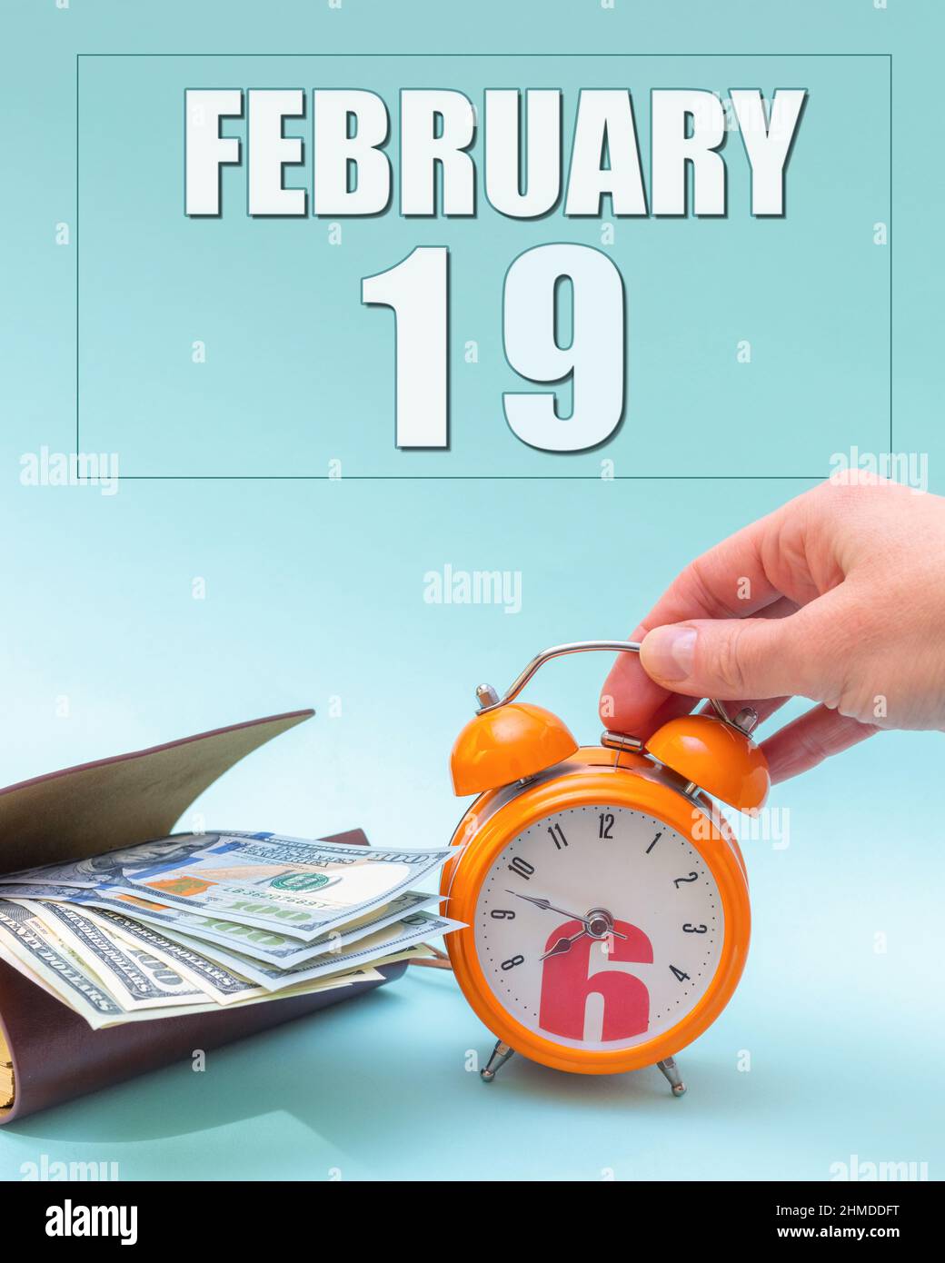 Febbraio 19th. Mano che tiene una sveglia arancione, un portafoglio con contanti e una data di calendario. Giorno 19 del mese. Pianificazione aziendale. Il tempo è denaro. Ora fiscale Foto Stock