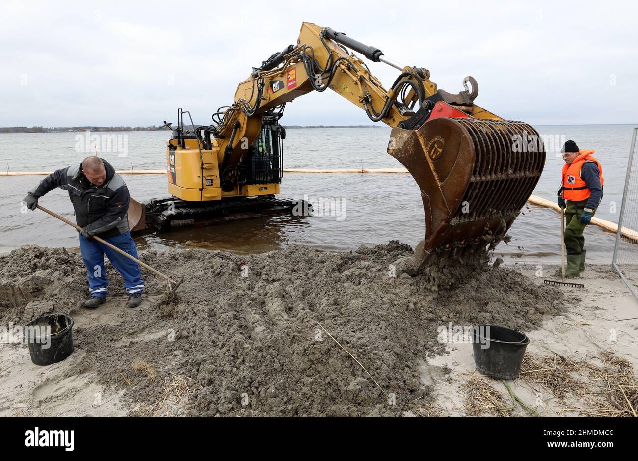 Rechlin, Germania. 09th Feb 2022. Presso la spiaggia di balneazione sul Müritz, un escavatore è utilizzato per rimuovere il terreno contaminato da pezzi di olio pesante dall'acqua; un braccio di petrolio protegge il sito. Circa 15 tonnellate di olio pesante sono dette per giacere come grumi catrame-come e zolle al fondo dell'acqua. Il sito contaminato risale al periodo in cui l'area fu utilizzata per scopi militari nel 1930s. Alcune parti della spiaggia di Müritz a Rechlin dovevano essere chiuse a causa dei grumi simili a catrame, che si sono lavati di nuovo e di nuovo. Credit: Bernd Wüstneck/dpa-Zentralbild/dpa/Alamy Live News Foto Stock