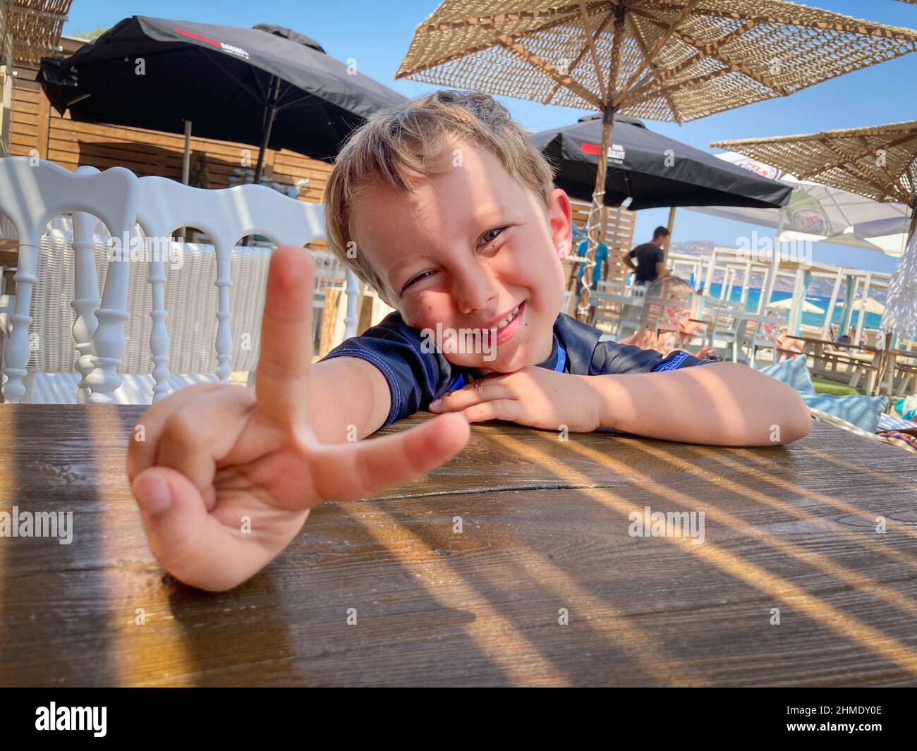 Ragazzo sorridente che mostra un cartello di pace mentre si siede sulla sedia al tavolo Foto Stock