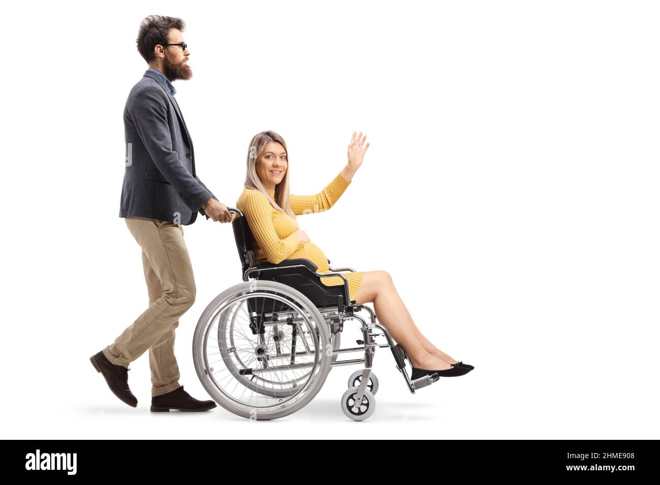 Colpo di profilo a tutta lunghezza di un uomo barbuto che spinge una donna incinta seduta su una sedia a rotelle e che agita isolato su sfondo bianco Foto Stock