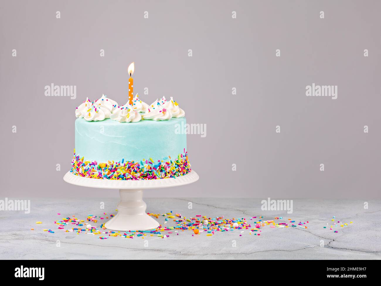 Torta di compleanno blu con spolverini colorati e candela di compleanno arancione illuminata su sfondo grigio chiaro. Foto Stock