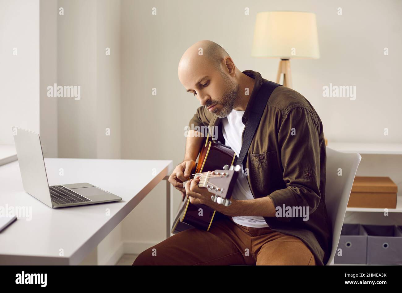 Uomo adulto focalizzato a casa esercitandosi a suonare la chitarra acustica mentre legge le note dal portatile. Foto Stock