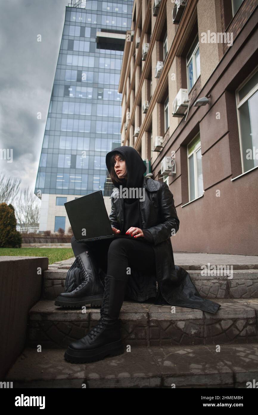 Ragazza in un cappotto di pelle nera e un cappuccio con computer portatile su una strada della città, concetto di una donna hacker e agente che lavora a distanza al computer Foto Stock