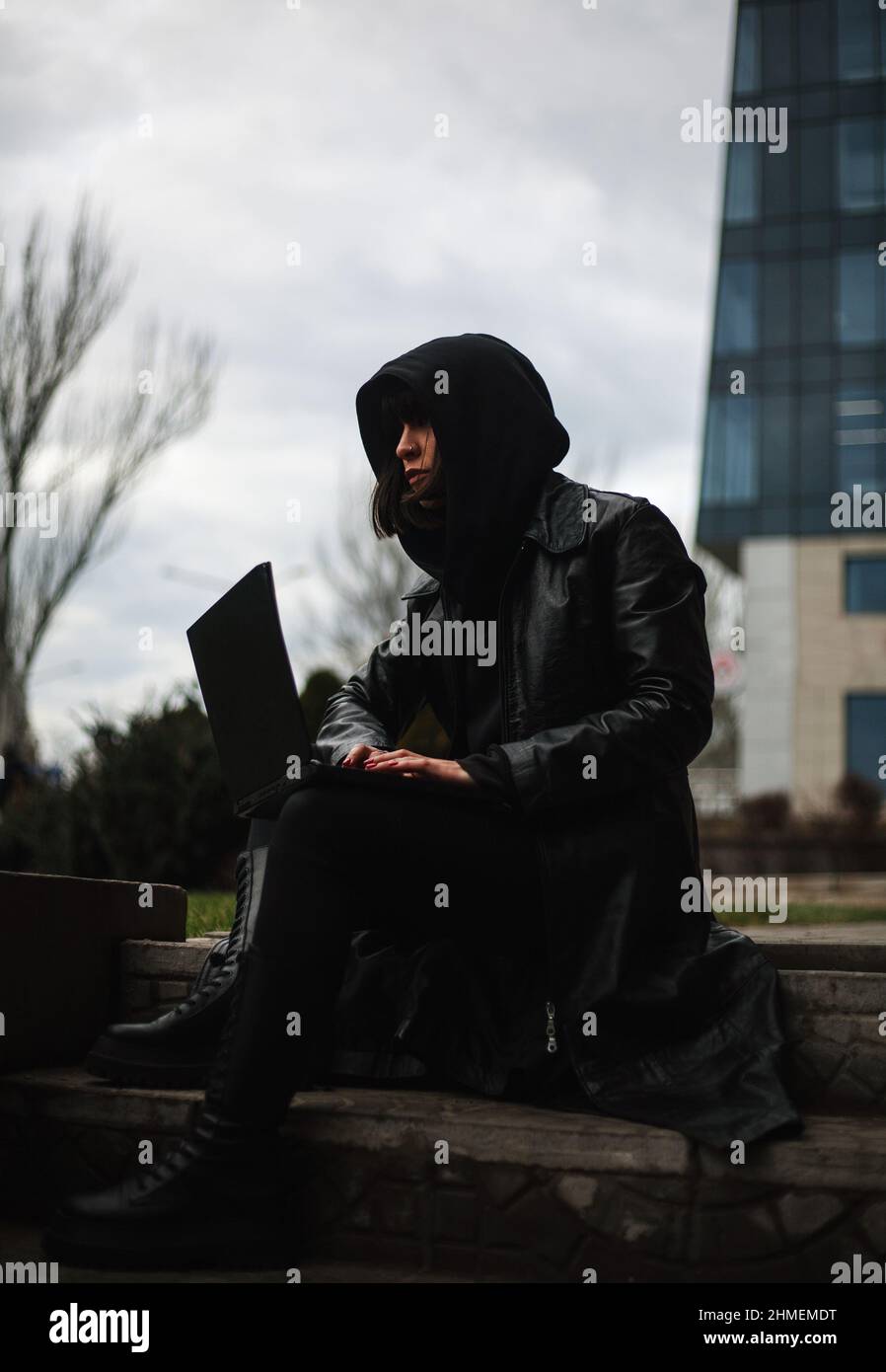 Ragazza in un cappotto di pelle nera e un cappuccio con computer portatile su una strada della città, concetto di una donna hacker e agente che lavora a distanza al computer Foto Stock