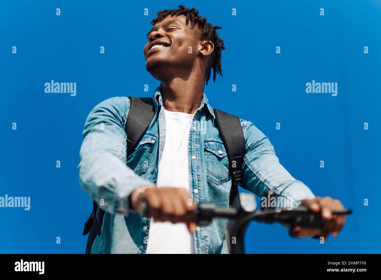 Felice sorridente uomo afroamericano ricurly in abiti casual che cavalca uno scooter elettrico in strada in un giorno di sole.gioioso bell'uomo a cavallo di un sco Foto Stock