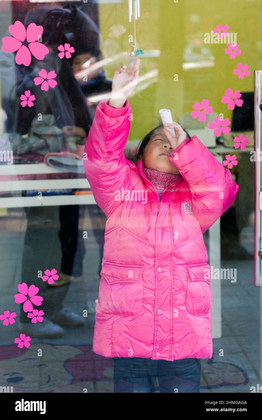 Giovane ragazza vestita in un anorak rosa che gioca dietro una porta di vetro decorata con fiori rosa. Jiashan, Cina Foto Stock