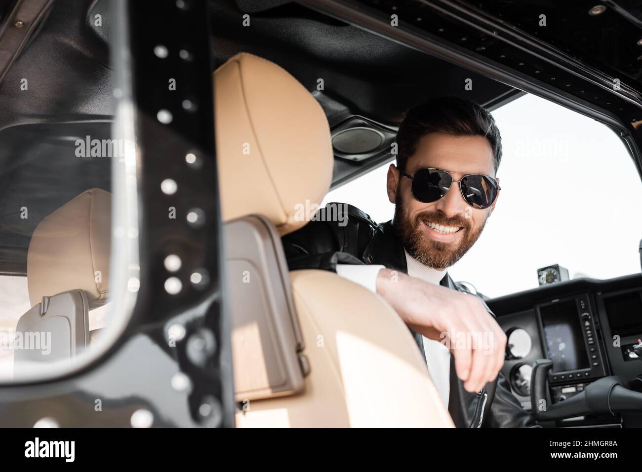 pilota positivo in eleganti occhiali da sole e giacca in pelle seduti nella cabina di pilotaggio di elicottero Foto Stock