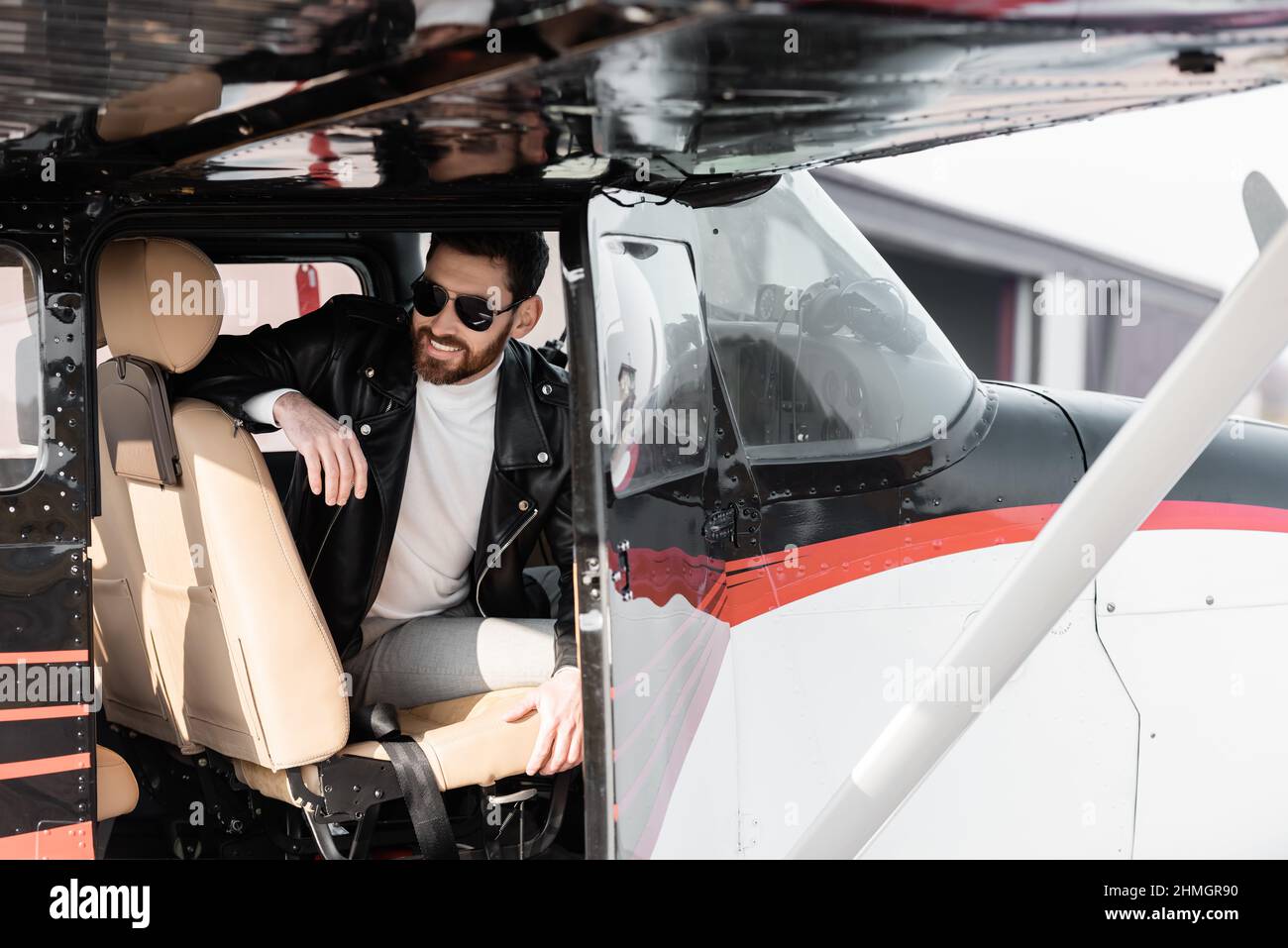 pilota bearded in occhiali da sole eleganti e giacca di pelle sorridente mentre si siede nella cabina di pilotaggio di elicottero Foto Stock