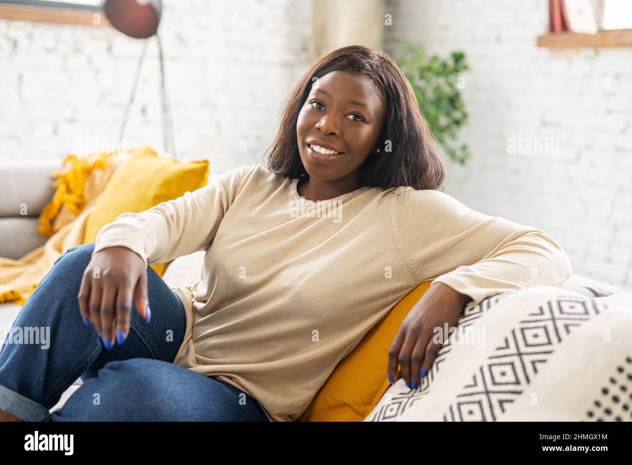 Affascinante donna afroamericana riposo a casa, serena etnia femminile seduta sul comodo divano in soggiorno moderno, sorridente e guardare la macchina fotografica. Donna calma e spensierata in abbigliamento casual Foto Stock